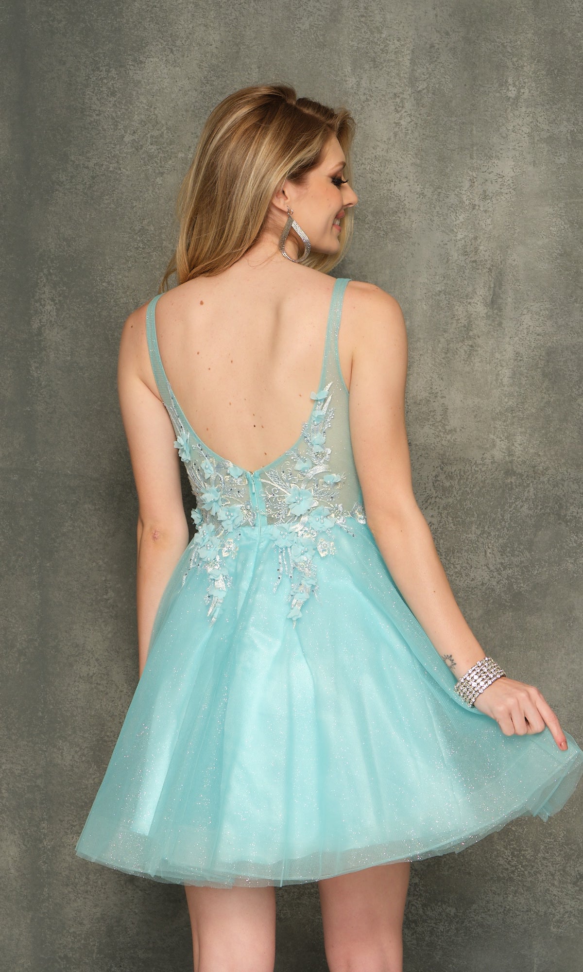 A woman faces away against a textured gray background, wearing the Sheer-Bodice Short Homecoming Dress in light blue with floral embellishments and a sleeveless design.